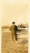 Image shows a black and white photograph of a person dressed in overalls and a toque standing outdoors. 