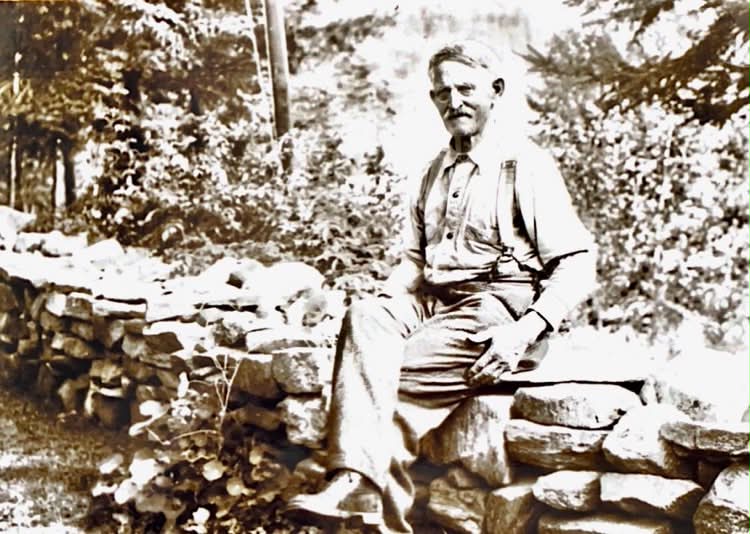 Image shows a black and white picture of a man sitting on a stone wall.