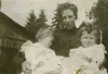 Image shows a woman holding two children in white gowns. 