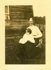 Image shows a black and white photo of a woman with a child on her lap seated outside a wooden building. 