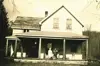 Image shows a black and white photo of a wooden farmhouse with people standing on the porch. 