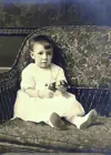 Image shows a small girl in a white dress seated on a sofa. 