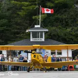 Image shows the Stanhope Municipal Airport terminal building with a crowd and yellow plane in the foreground. 