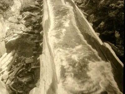 Image shows a black and white photo of the log chute with water flowing through it. 